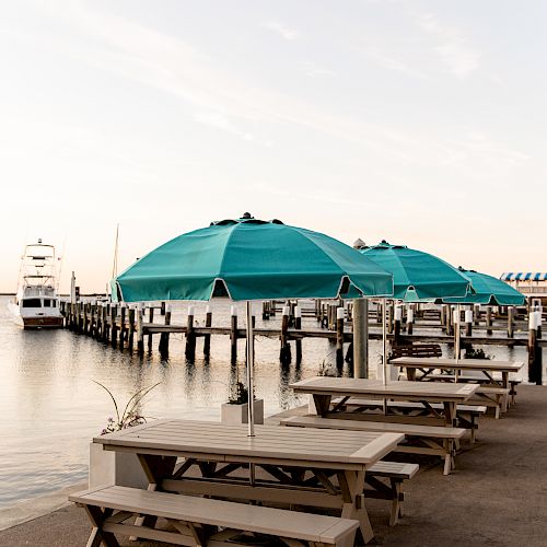 The image shows picnic tables with teal umbrellas by a dock on a calm body of water, with a boat and partially visible structures in the background.