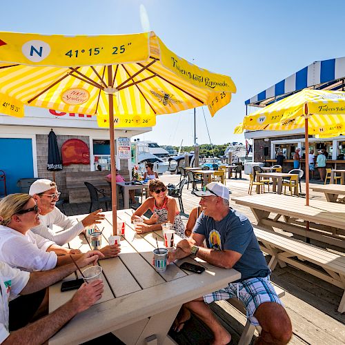 People sitting at outdoor picnic tables under yellow umbrellas, enjoying drinks and conversation. The location appears to be a beachside cafe.