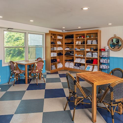 A cozy room with checkered flooring, tables, chairs, shelves of clothing, towels, and toiletries, plus a round mirror and wall-mounted items.