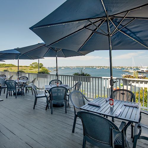 Outdoor dining area with tables, chairs, and large umbrellas on a wooden deck overlooking boats and water under a clear blue sky.