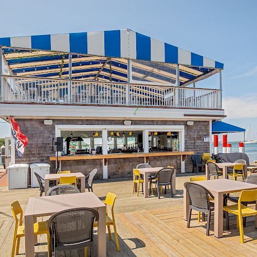 An outdoor waterfront restaurant with yellow and gray chairs, wooden tables, and a striped blue-and-white canopy. Nearby, boats are docked.