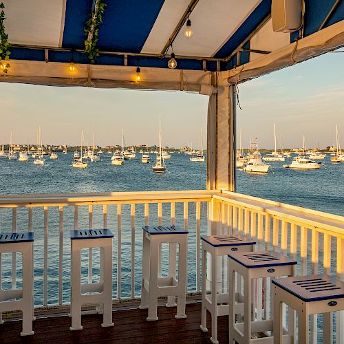 Outdoor seating area with bar stools and high tables overlooking a marina with numerous boats on the water during the day, under a blue and white canopy.