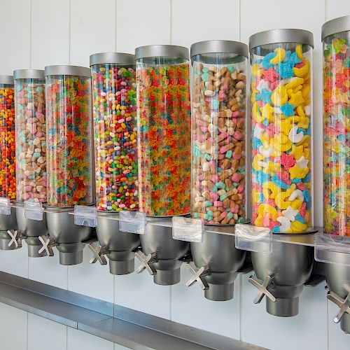 The image shows a row of clear dispensers attached to a wall, filled with various colorful candies, such as gummies and jelly beans.