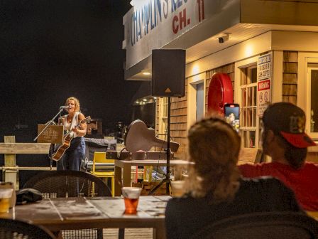 A musician performs on a dockside stage at night while a couple watches from a table with drinks, under a building with 