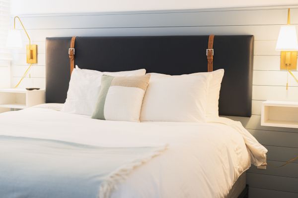 A neatly made bed with a padded headboard, white linens, and several pillows, flanked by wall-mounted bedside tables and reading lamps.