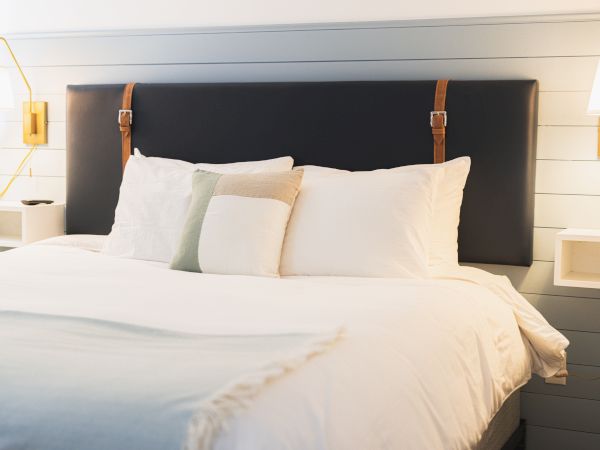 A neatly made bed with a padded headboard, white linens, and several pillows, flanked by wall-mounted bedside tables and reading lamps.
