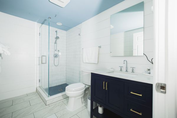 The image shows a modern bathroom with a glass-enclosed shower, a toilet, and a dark vanity with a white countertop and mirror, all in a bright space.