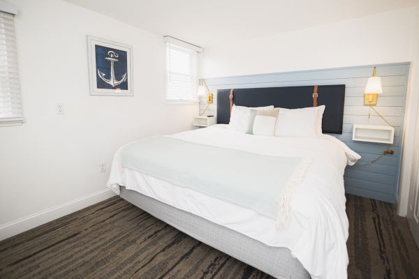 A neatly arranged bedroom with a large bed, white linens, a nautical-themed painting, and wall-mounted lamps on either side of the bed.