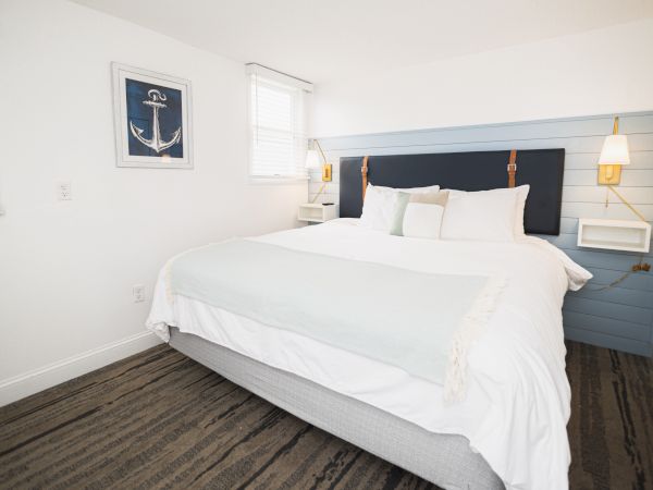 A neatly arranged bedroom with a large bed, white linens, a nautical-themed painting, and wall-mounted lamps on either side of the bed.
