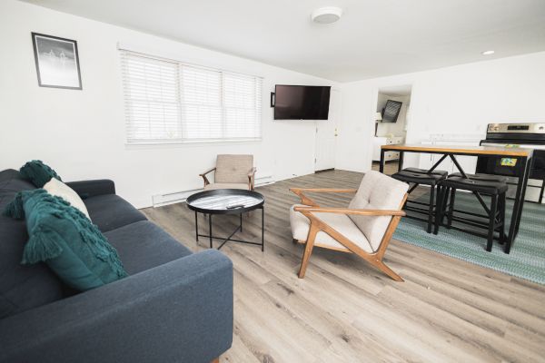 A modern living area with a sofa, two chairs, a round coffee table, a TV, and a kitchen area with countertops and bar stools in the background.