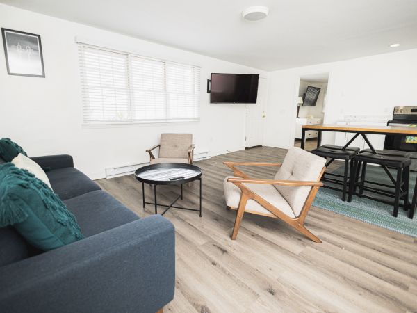 A modern living area with a sofa, two chairs, a round coffee table, a TV, and a kitchen area with countertops and bar stools in the background.