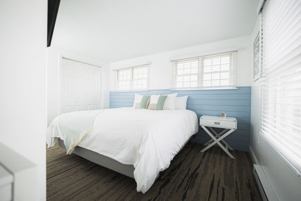 A bright and modern bedroom with a large bed, white and pastel decor, a blue accent wall, multiple windows, and a side table with a clock.