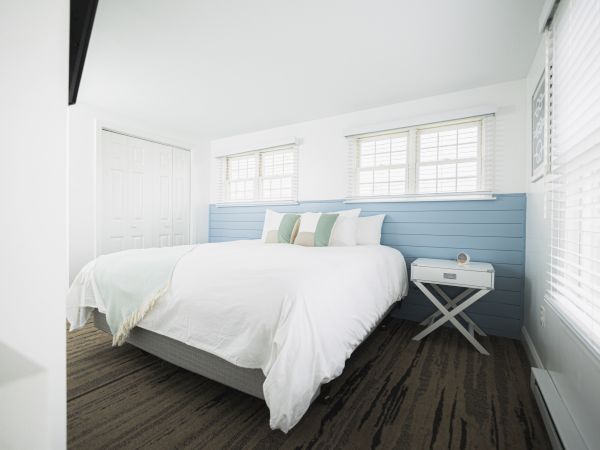 A bright and modern bedroom with a large bed, white and pastel decor, a blue accent wall, multiple windows, and a side table with a clock.