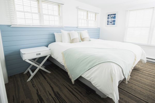 The image shows a clean, minimalistic bedroom with a large bed, white and light blue bedding, and a small side table on a wood-patterned floor.