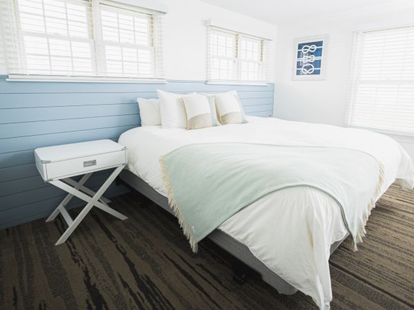 The image shows a clean, minimalistic bedroom with a large bed, white and light blue bedding, and a small side table on a wood-patterned floor.