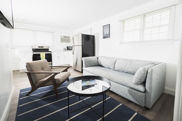 A modern living room with a gray sofa, armchair, round glass-top table, blue striped rug, and a view into an open kitchen.