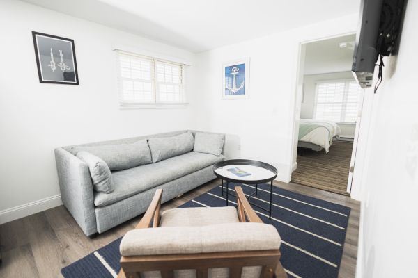 This image shows a minimalist living room with a gray couch, a wooden armchair, a round black table, and nautical-themed decor. Bedroom visible.