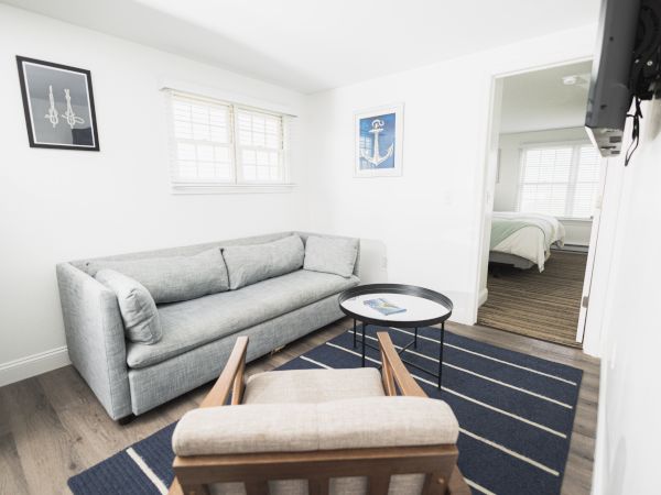This image shows a minimalist living room with a gray couch, a wooden armchair, a round black table, and nautical-themed decor. Bedroom visible.