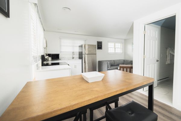 A modern kitchen and living area with white walls, wooden table, black chairs, stainless steel appliances, and a gray sofa beside a window.