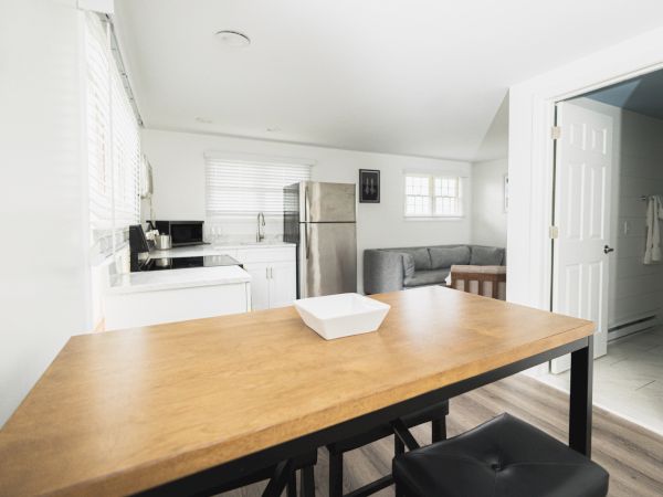 A modern kitchen and living area with white walls, wooden table, black chairs, stainless steel appliances, and a gray sofa beside a window.