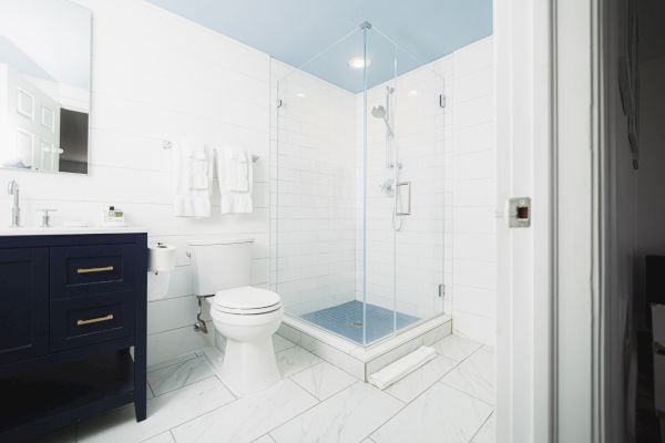 A modern bathroom features a glass-enclosed shower, white toilet, black vanity with a mirror, and white tiled walls and floor, with towels hanging.