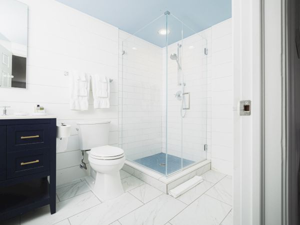 A modern bathroom features a glass-enclosed shower, white toilet, black vanity with a mirror, and white tiled walls and floor, with towels hanging.