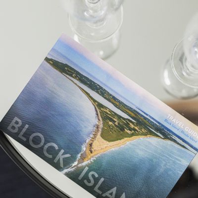 The image shows a postcard placed on a glass surface. The postcard features an aerial view of Block Island, a coastal area with sandy shores.