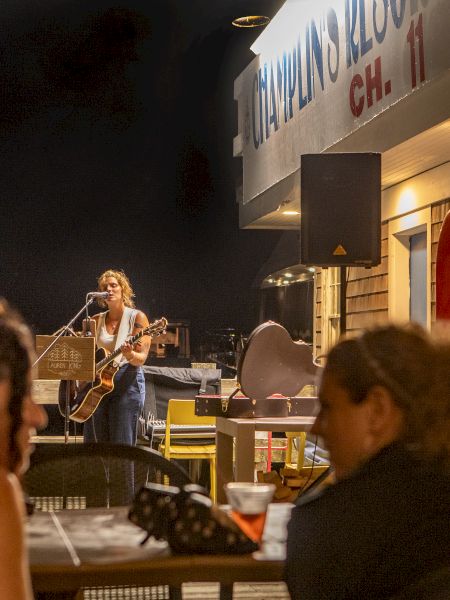 A musician performs on an outdoor stage at night, while a few people sit at tables enjoying drinks and watching the performance.