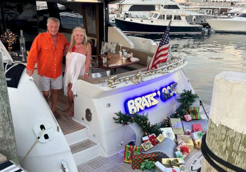 Two people stand on a yacht decorated with gifts and an American flag, docked at a marina, with presents arranged at the stern.