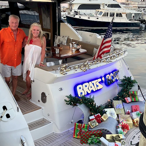Two people stand on a yacht decorated with gifts and an American flag, docked at a marina, with presents arranged at the stern.