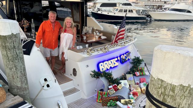 Two people stand on a yacht decorated with gifts and an American flag, docked at a marina, with presents arranged at the stern.