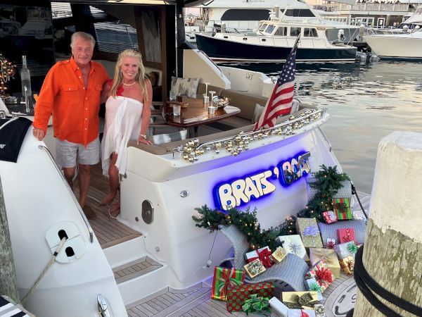 Two people stand on a white boat loaded with wrapped gifts and a festive setup, docked at a marina with other boats nearby, American flag fluttering.
