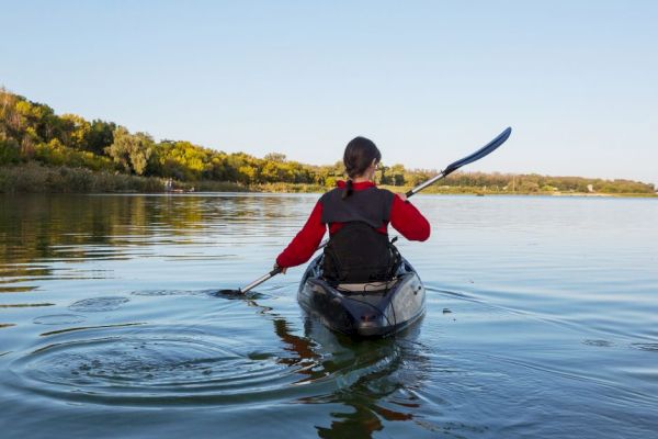 Someone kayaking on a calm lake, surrounded by trees and under a clear sky. The person is wearing a red jacket and a life vest.