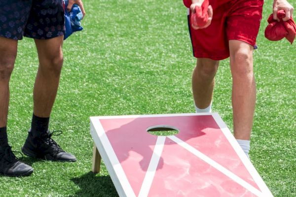 Two people are playing cornhole on a grass field. One is throwing a bag while the other holds blue bags.