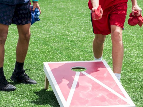 Two people are playing cornhole on a grass field. One is throwing a bag while the other holds blue bags.