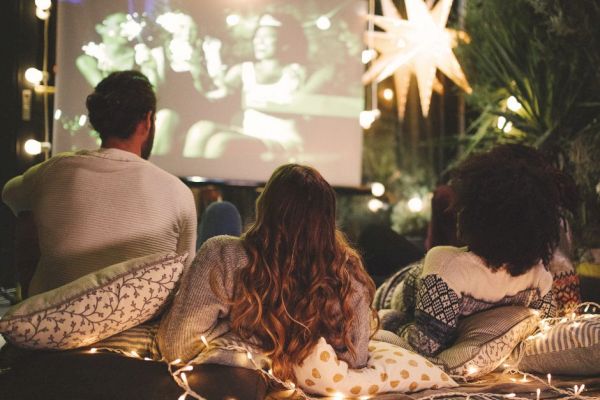 Three people are sitting on pillows outdoors, watching a movie on a large screen surrounded by string lights and a glowing star decoration.