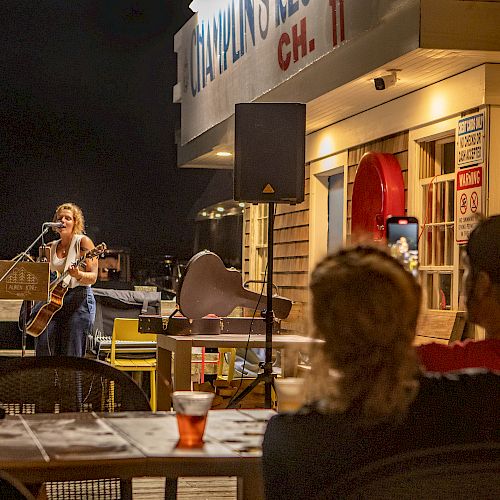 A musician performs with a guitar in an outdoor venue at night, while an audience watches from tables with drinks and a guitar case nearby.