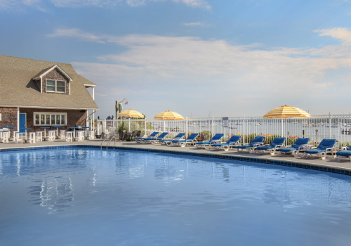 This image shows an outdoor pool area with a house-like building, numerous lounge chairs, and yellow umbrellas overlooking a body of water.