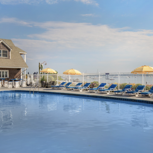 This image shows an outdoor pool area with a house-like building, numerous lounge chairs, and yellow umbrellas overlooking a body of water.