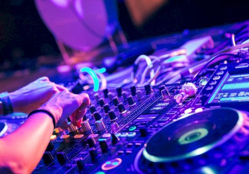 A DJ is mixing music on a console with various controls and turntables, surrounded by colorful lights, possibly at a club or event.