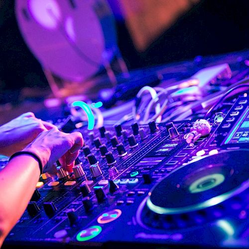 A DJ is mixing music on a console with various controls and turntables, surrounded by colorful lights, possibly at a club or event.