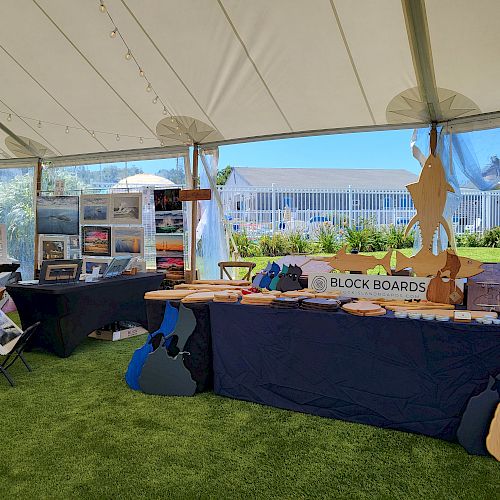 The image shows an outdoor exhibition booth with surfboards, displayed artwork, and other items under a tent on a grassy area, near a white fence ending the sentence.