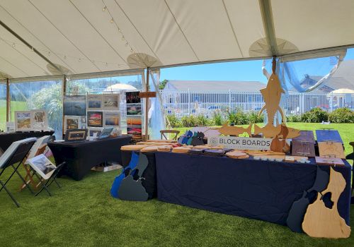 The image shows a sunny art fair with various booths displaying colorful artwork and handcrafted items, set up under a white tent with open sides.