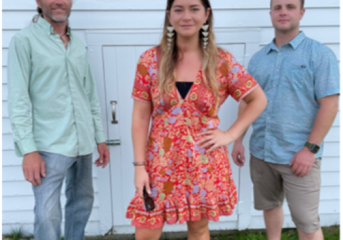 Three individuals stand in front of a white wall with the text "Nena & The Cosmos" above them. The woman in the center wears a floral dress.