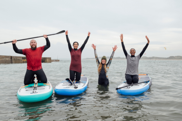 Four people are enjoying paddleboarding; three standing on boards and one in the water. They have their arms raised in excitement.