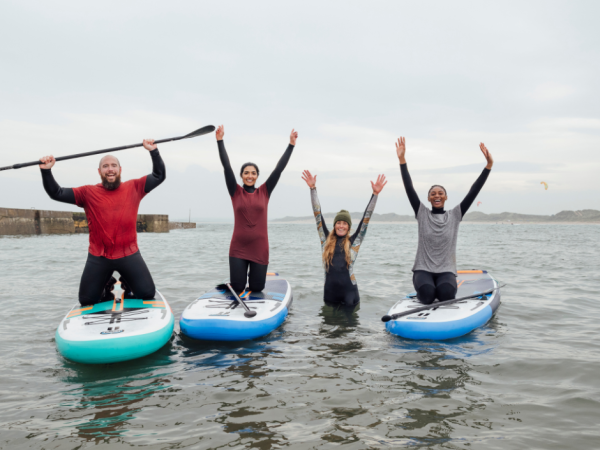 Four people are enjoying paddleboarding; three standing on boards and one in the water. They have their arms raised in excitement.