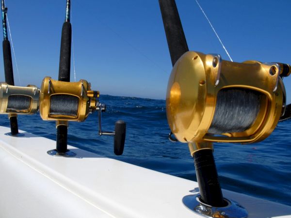 Three fishing reels mounted on a boat rail, ready for offshore fishing, with blue ocean and clear sky in the background.