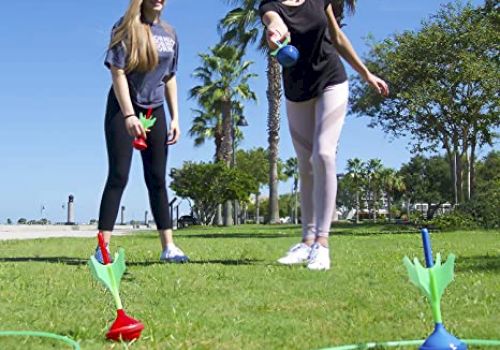Two people are playing an outdoor lawn game, tossing weighted darts into rings on the ground with trees and a blue sky in the background.