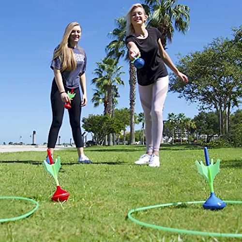 Two people are playing an outdoor lawn game, tossing weighted darts into rings on the ground with trees and a blue sky in the background.