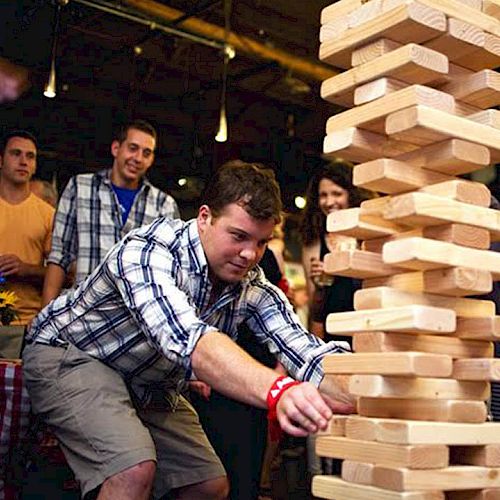 A group of people is playing a giant Jenga game in what appears to be a social gathering, with a man carefully pulling out a block.